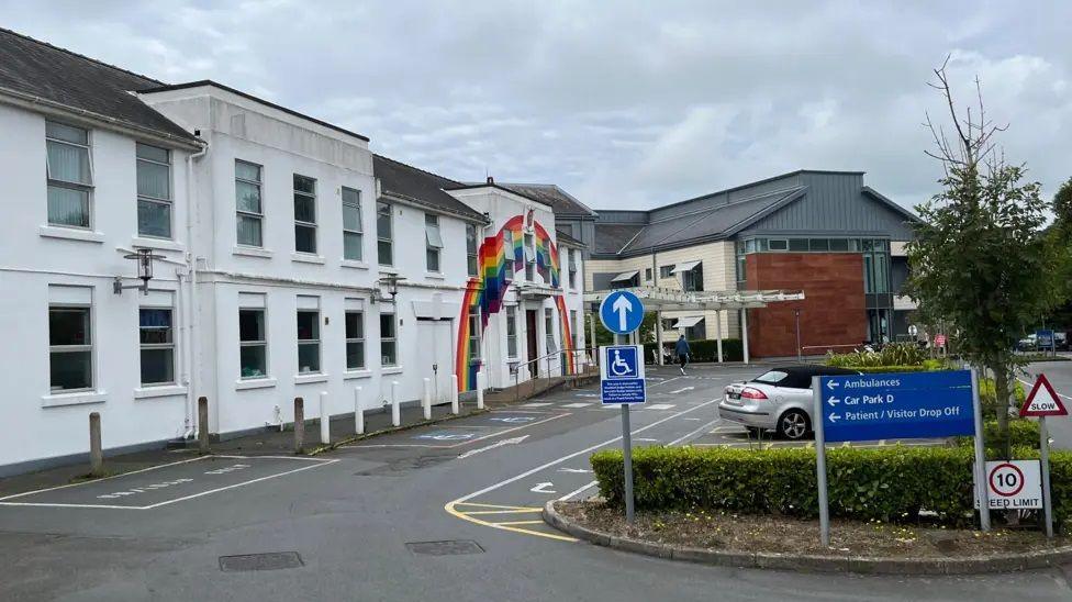 Photo of the outside of the Princess Elizabeth Hospital. A rainbow has been painted across the white building. The car park has one car, a tree and a hedge, plus a couple blue signs.