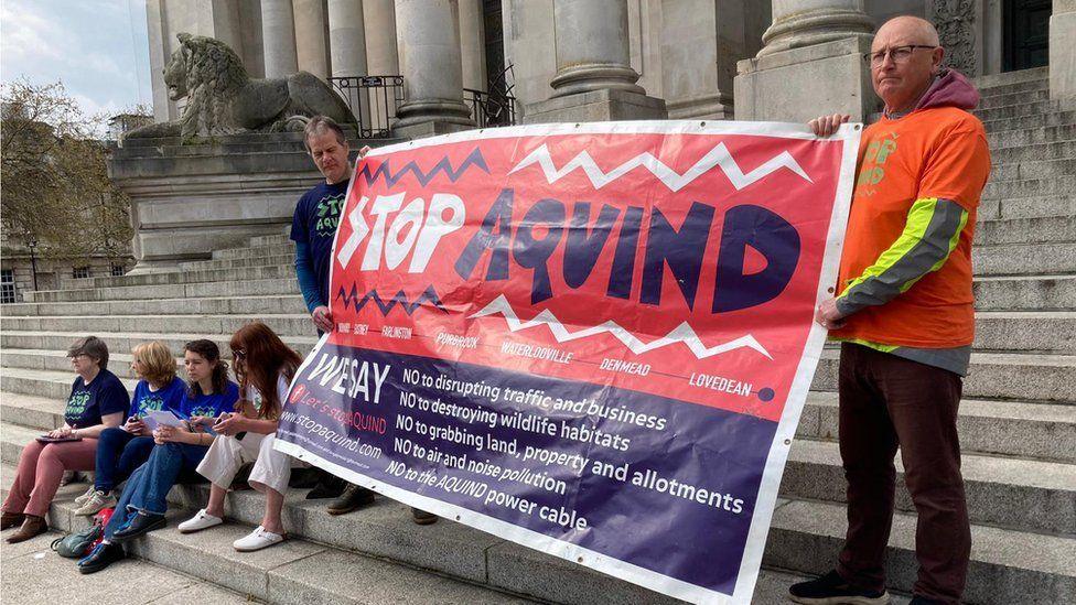 Protestors on the steps of a civic building in Portsmouth hold a large banner with the words "Stop Aquind"