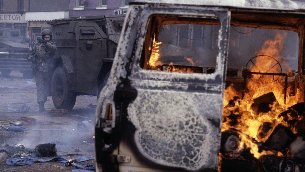 A car on fire with army personnel standing behind it during the Troubles.