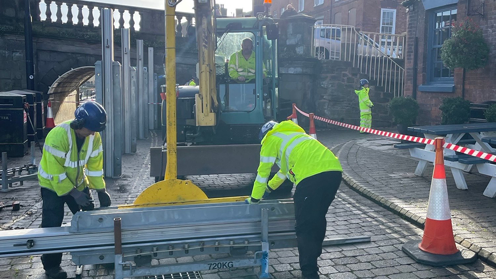Two men in blue hard hats and high-vis jackets putting up flood defences, while another is in a mini digger lifting metal parts in to the pavement. An arch of a bridge is in the background
