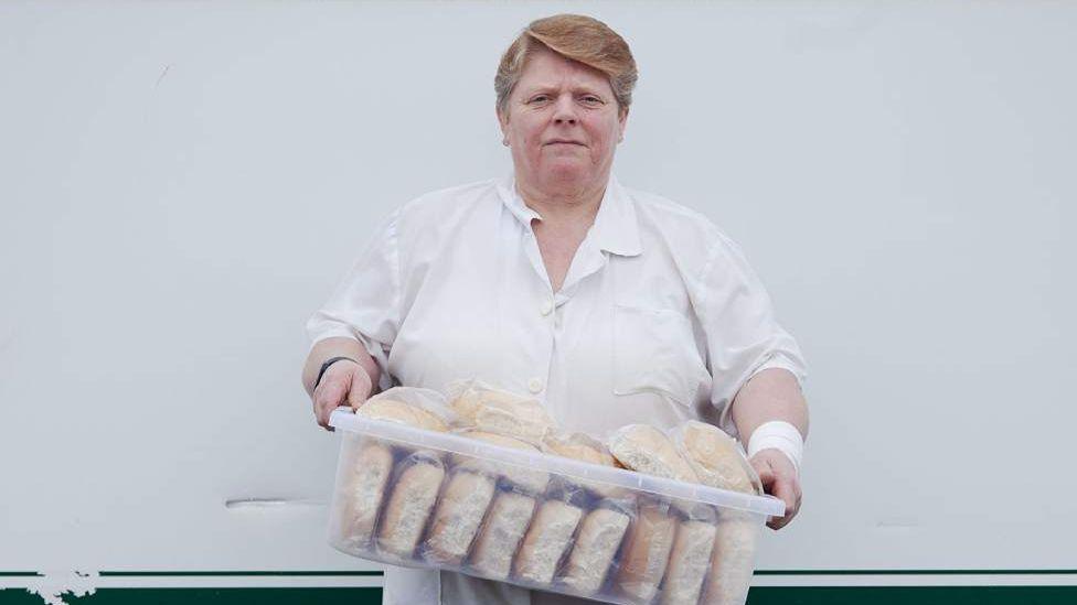 A woman dressed in white overalls holds a large plastic container holding bread rolls for her burger van used by steelworkers