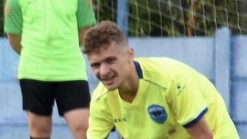 Kyle Shimwell is pictured wearing a fluorescent yellow football shirt. A goal net is in the background. He has curly short hair.