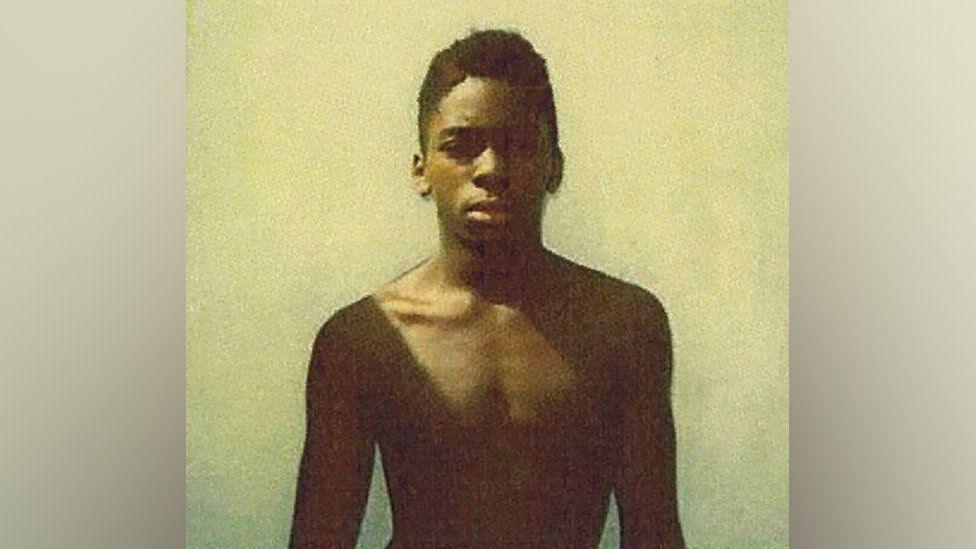 A police image of Mark Gordon taken at the time of his arrest in 1989. He is standing bare-chested against a yellow background and is looking directly ahead.
