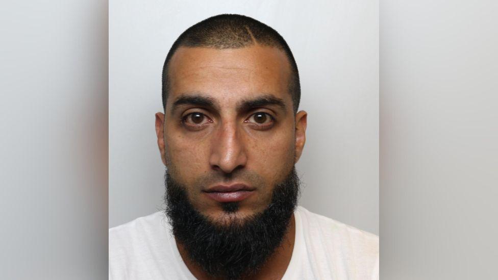 The mugshot of a man with shaved dark hair and a large beard. He is wearing a white t-shirt and looking directly and solemnly into the camera.