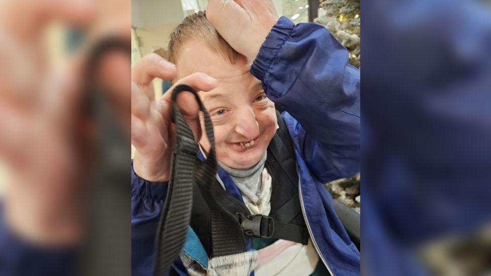 Ryan wearing a blue rain jacket and a black chest strap with buckles. He is smiling and looks excited and happy with both his hands in the air.