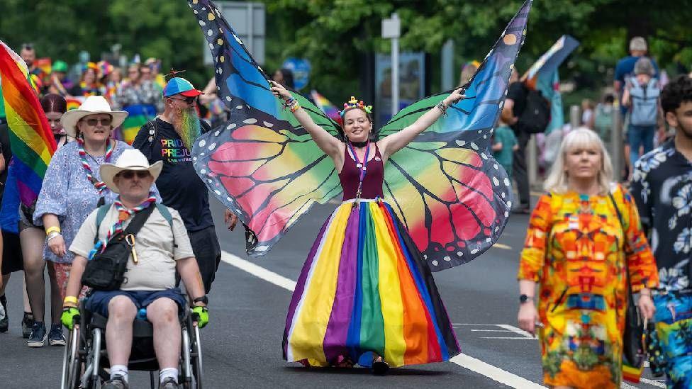 A person dressed as a buttefly, wearing a red vest top and a rainbow-coloured skirt.