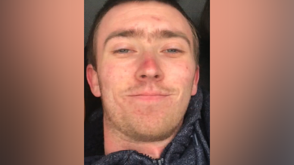 A selfie of Daniel Clarke. He has short, light brown hair and stubble, and is wearing a dark grey hoodie as he smiles at the camera