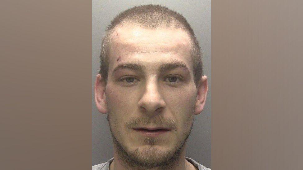 A police mugshot of Jonathon Newby. He has a short moustache and beard and short brown hair.