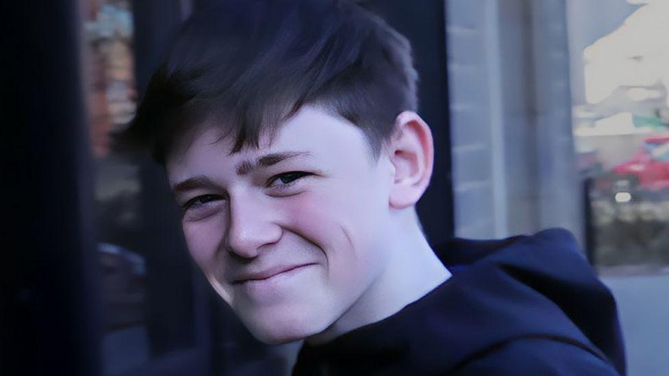 A boy with brow hair, wearing a navy hoodie, smiles at the camera, standing outside.