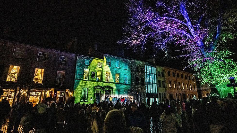 A light projection  in green colours on buildings on Dalton Square in the dark, with a lit up tree to the right and lots of people milling about in front of the buildings