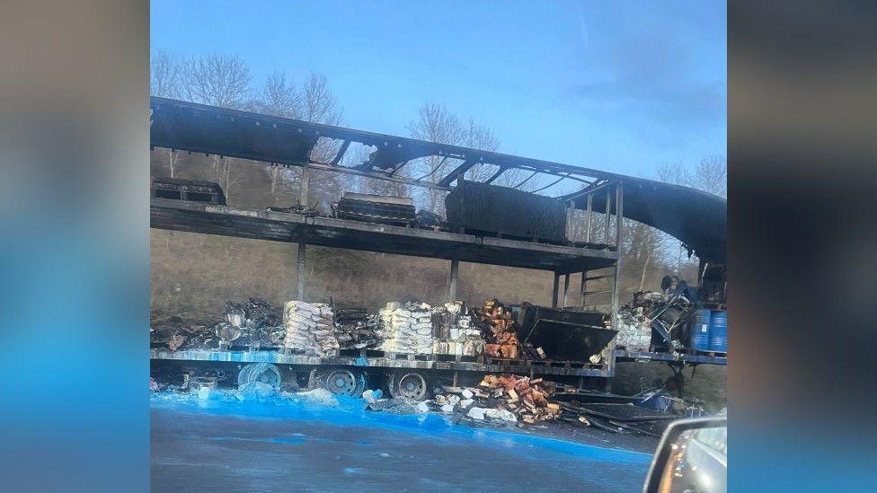 The aftermath of a lorry fire on the M40 motorway. The picture shows a view of the burnt-out lorry from a car window. The outer parts of the vehicle are burnt away, leaving only parts of the structure, the wheels, and the goods the lorry was carrying. Some of them have toppled onto the round and many of them are visibly burnt. A car's wing mirror is visible in the bottom right corner of the photo.