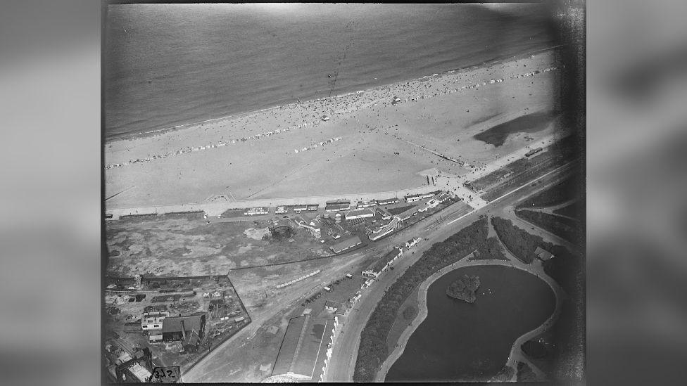 A black-and-white aerial shot of a road and a beach with lots of people on it.