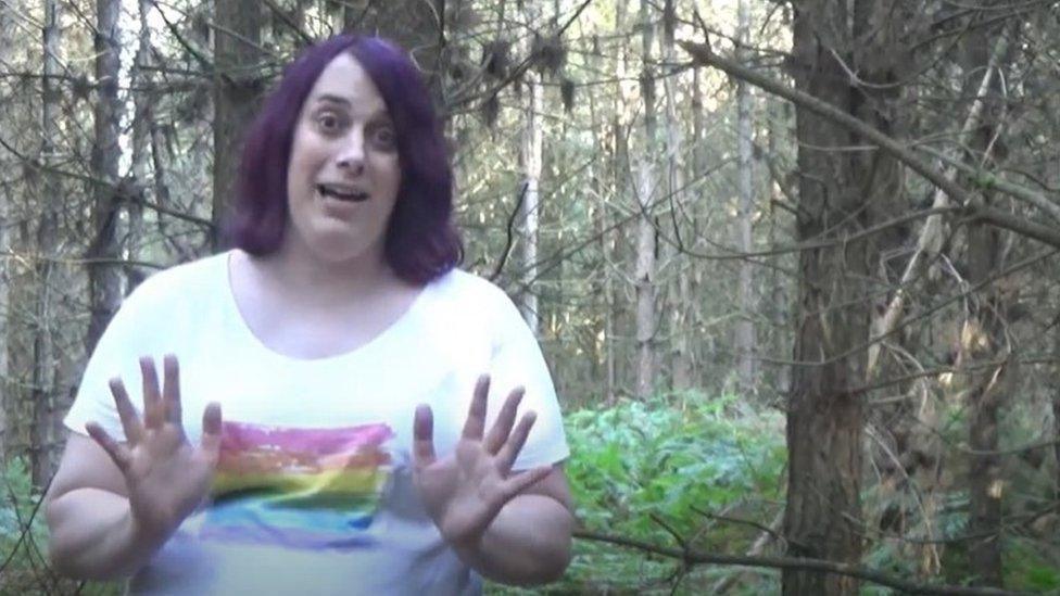 Zoe Watts stands in a forest surrounded by trees. She has shoulder length purple hair and is wearing a white T-shirt with a rainbow coloured square on it. Her mouth is open and she has both hands held out in front of her.