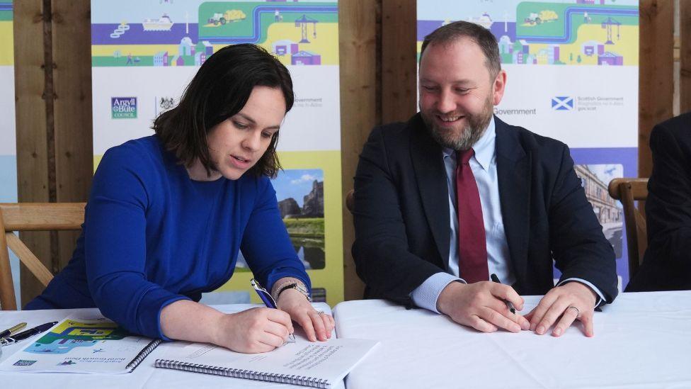 Forbes and Murray sit at a table, with Forbes singing a document. She is wearing a blue dress and looking down at the document. On her left Murray, wearing a dark suit and red tie, is smiling with a pen in his hand, also looking down at the document. 