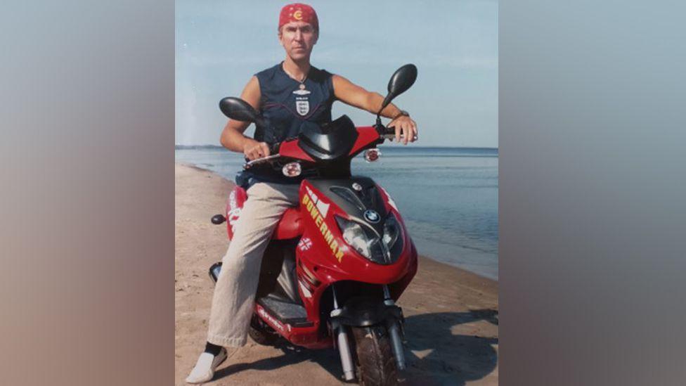 Maris Ile is sitting on a red motorbike next to the sea on a beach. He is wearing a sleeveless blue top and beige trousers. He is also wearing a red bandana on his head and white pumps on his feet. 