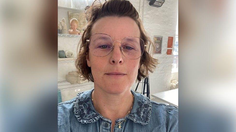 A photo showing the face of a woman wearing glasses. She has brown hair and is stood in front of a white wall and shelves of arts and crafts