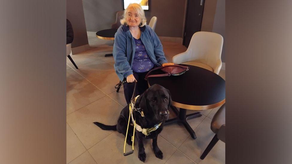 Kim Milward and her guide dog Woody, a black labrador, sit in a cafe.
