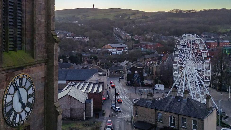 Ariel view of Darwen town centre with the observation wheel to the right, Darwen Tower and a church clock tower to the left, and the moors in the distance.