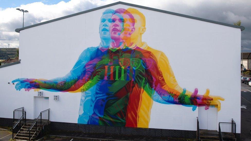 A mural on a white wall of footballer James McClean - he is wearing a Republic of Ireland kit and his his arms outstretched