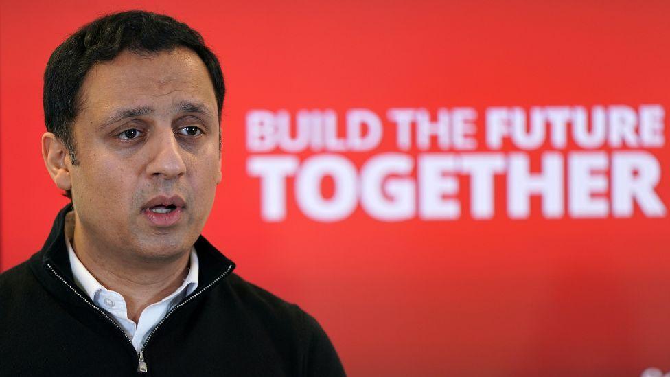 A man with dark hair, wearing a black jumper over a white shirt, speaks in front of a red Labour background in a medium close-up shot 