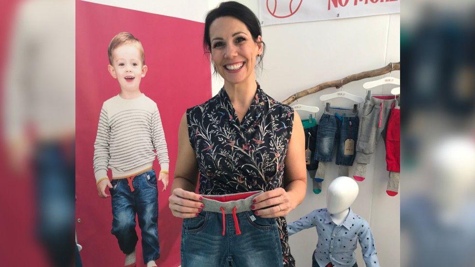 Natalie Hull standing in front of a Sockatoos advert in a black top and holding a pair of the sockatoos trousers