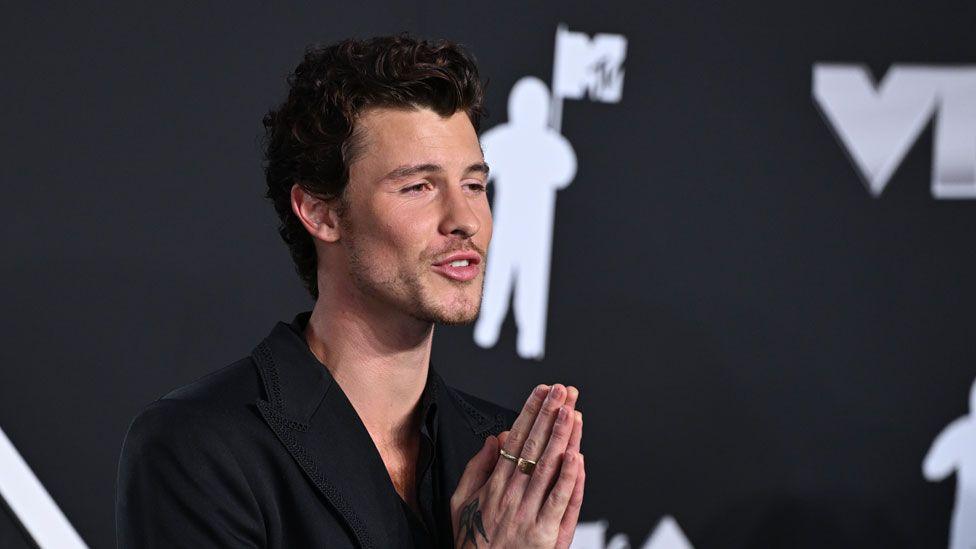 Shawn Mendes who has dark fairly short wavy hair and stubble. He is wearing a black jacket and looking towards his left with his hands up in prayer