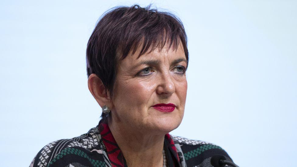 Angela Constance, a woman with short dark hair, photographed in a medium-close up. She is wearing a green, black and white patterned top. 