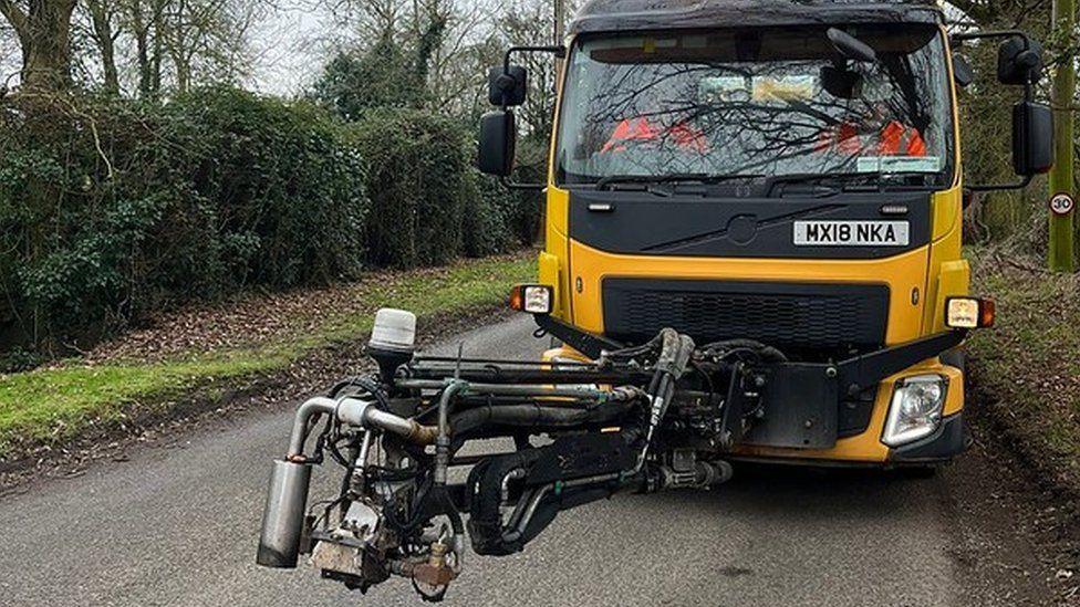 The Dragon Patcher machine is pictured on a rural road. It is a yellow and black lorry with an attached placed on the road with various different parts. The road is enclosed by bushes on either side.