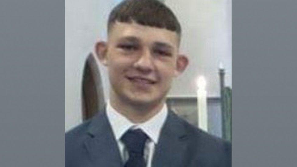 Nojukas Korzeniauskas, a young man wearing a grey jacket over a white shirt and dark tie. He has short dark hair cut in a short fringe over his forehead and shaved at the sides. Behind him can be seen a lit candle.