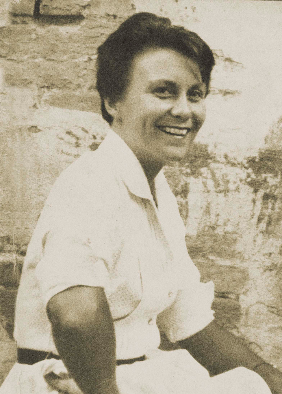 Sepia photo of a smiling Harper Lee looking to camera -- side on. with her right arm on her waist. She's wearing a white short sleeved blouse and white skirt.
Background og brick wall.