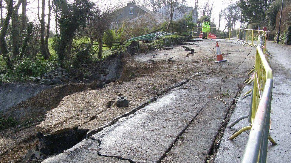 Undercliff Drive, with large cracks and holes in the tarmac. The area is fenced off and a policeman is on duty in the background.