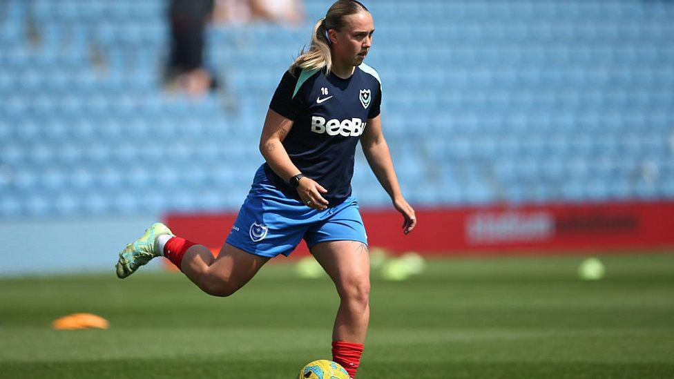 Ella Humphrey playing a game at Portsmouth FC Women. She is on the football pitch running and about to kick a ball. In the background are empty seats.