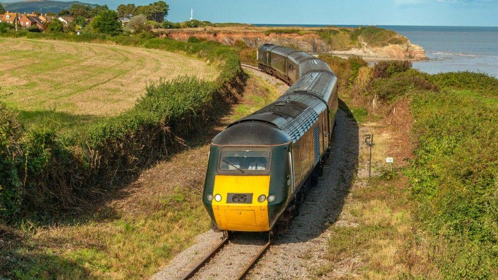A dark green train with a yellow front is travelling along a track next to the sea. It is sunny, the sky is blue.