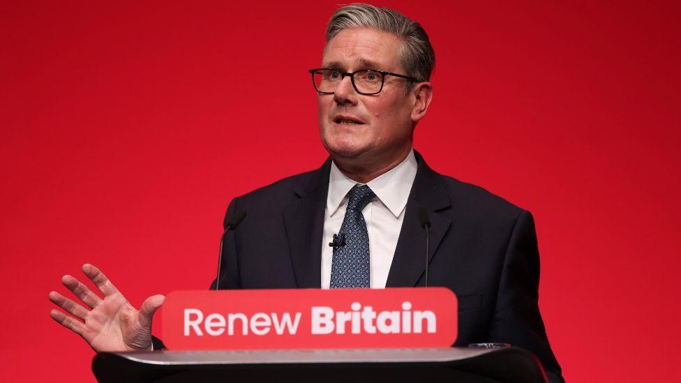 Keir Starmer, who has grey hair and glasses, speaks at a podium. He is wearing a dark suit, white shirt and blue tie. The background is red and there is a "renew Britain" sign in the foreground.