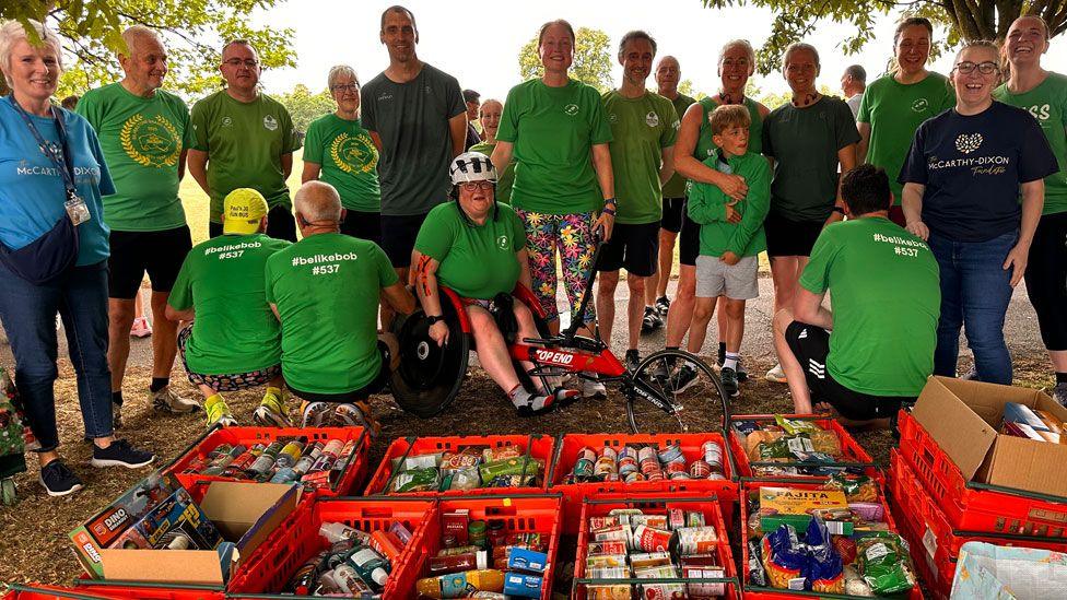 About 20 men and women of a range of ages, and a boy, standing behind red plastic crates filled with tins and packets of non-perishable food. Most are wearing green T-shirts, and those with their backs to us have #belikebob written on them in white letters. They are standing on parched, yellow grass. Behind them is sky and leafy tree branches.