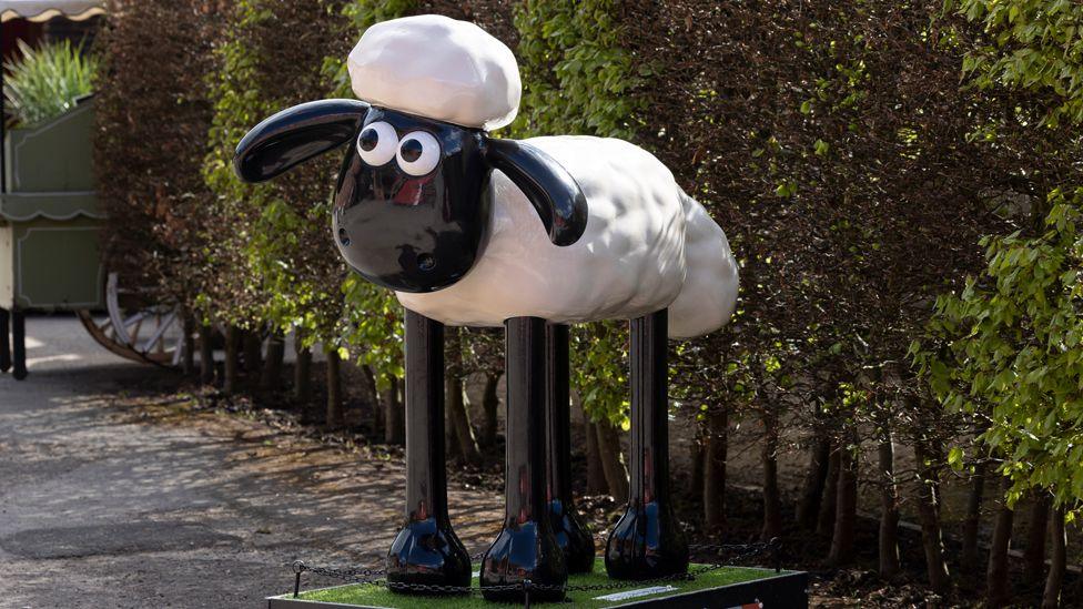 Model of character Shaun the Sheep on a plinth in front of a hedge. He has a black face and big black and white eyes, a white body and long black legs.