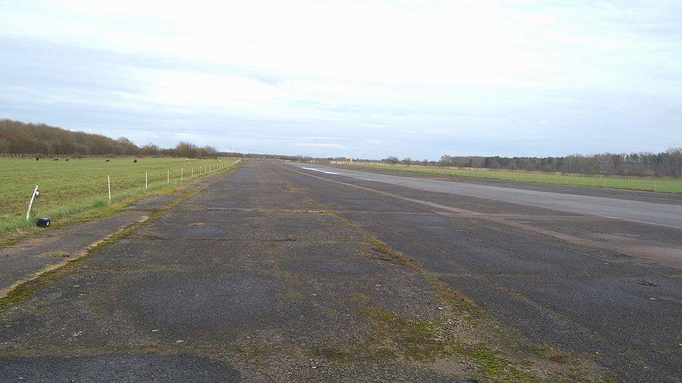The moss-covered black tarmac runway at Finmere Airfield. On either side are wide green fields, edged by trees.