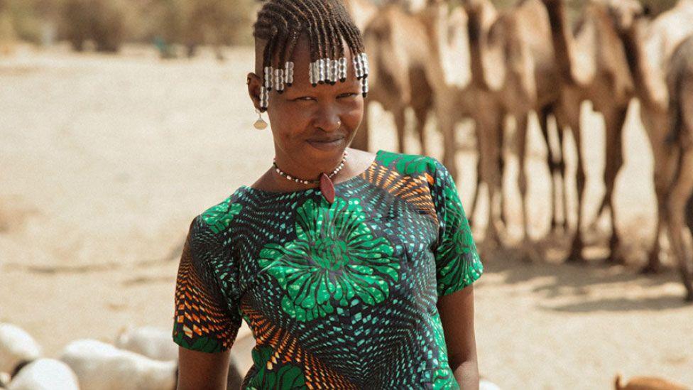 Apuu Mourrine wearing a patterned green top, bead necklace and beads in her braided hair smiles at the camera. Behind her can be seen camels and other livestock