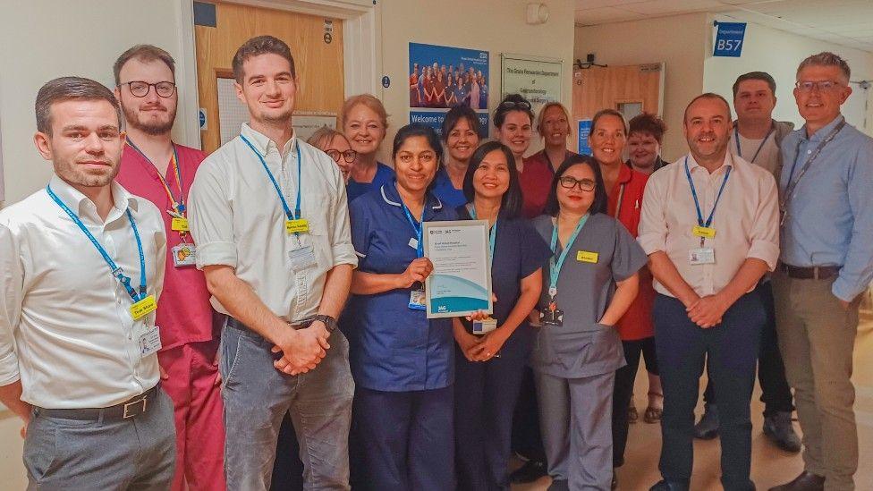 A group photo of people in shirts and scrubs on a hospital ward holding up a paper award. They are all smiling.