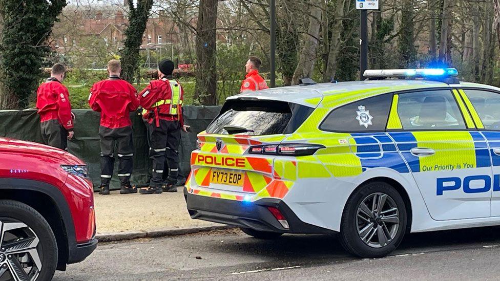 Four men in bright red jackets looking at the river, with a police car in front