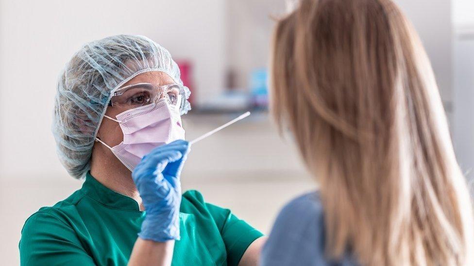 A clinician in green surgical gown, face mask, blue gloves and hair net holds a swab in front of a woman,who has blonde hair and who is standing directly opposite her