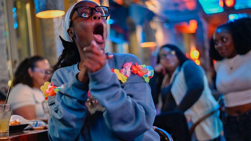 A woman at a Love Island USA watch party in Boston wearing a blue top with a lay of plastic flowers around her clasps her hands and is open-mouthed as she stares at a screen - July 2025.