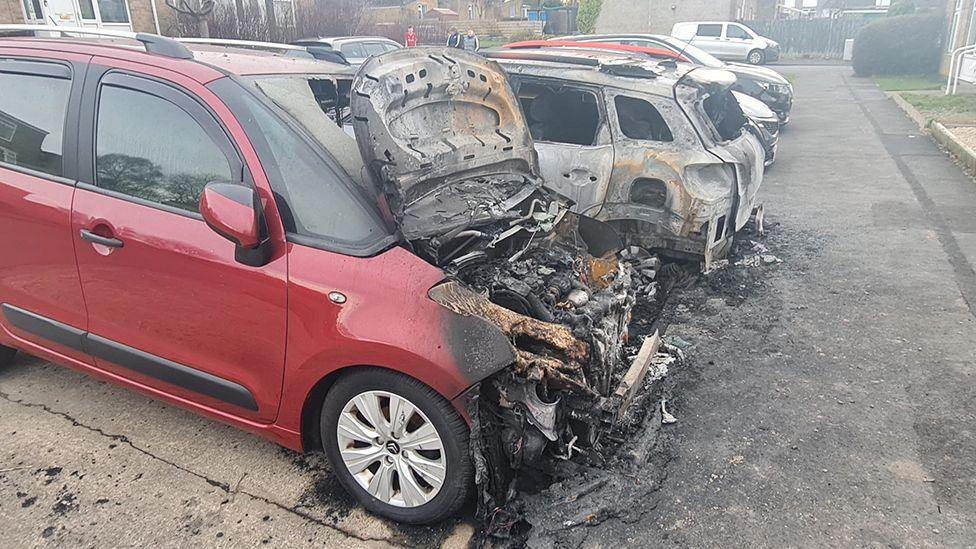 Two cars in a row of parked cars are severely burnt. The nearest one, which is red, has its bonnet open and the engine compartment is a mass of grey, tangled, burnt metal and charred remains. Beside it is a white car which is also burnt but is damaged less.