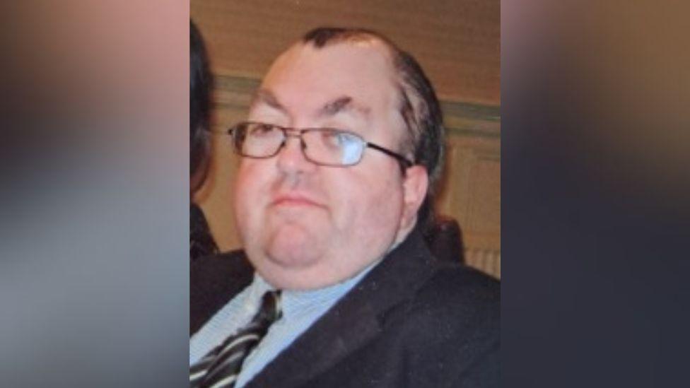 Graham Milliken looking straight at the camera. He has short, receding dark hair  and glasses. He is wearing a suit jacket with a blue shirt and a striped tie