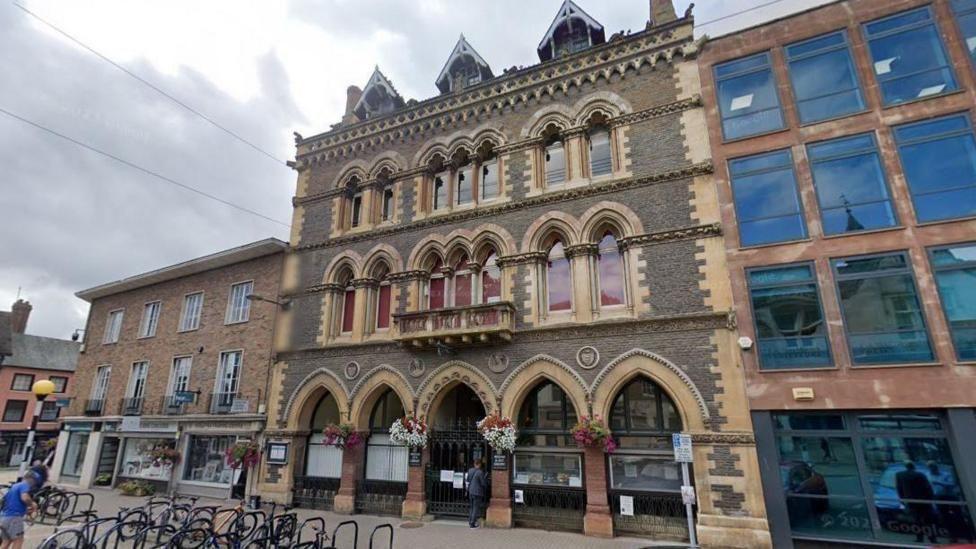 A historical three storey building made of brown bricks with a sandstone edge. On the ground floor it has five arches, which have flowers attached to their columns