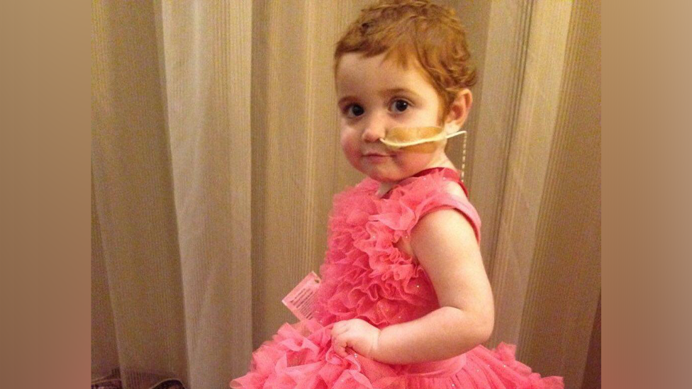 A toddler with red hair and a tube taped to her cheek, going into her nose, looks at the camera. She is wearing a pink frilly dress