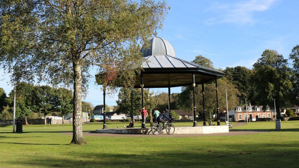A bandstand in green parkland. There are a few trees and some children playing on their bikes.