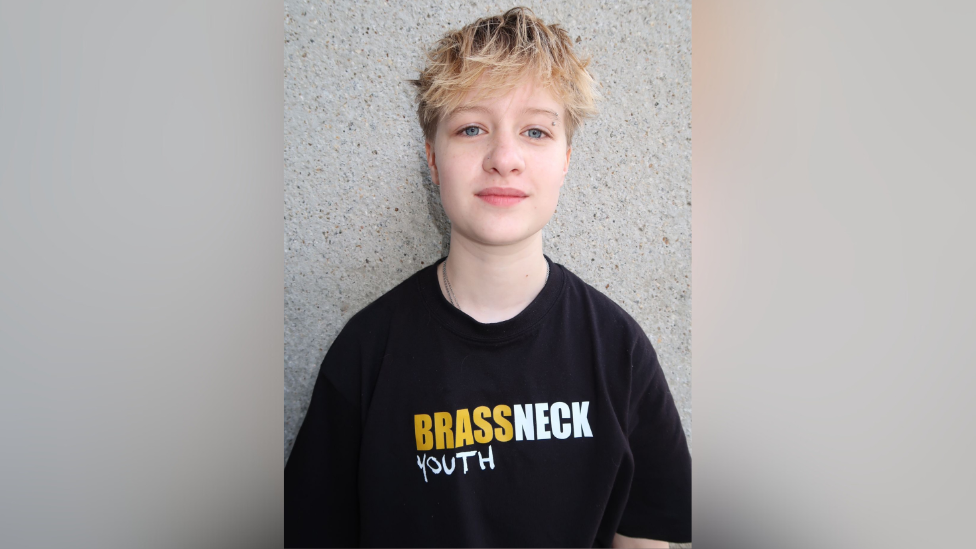 Fionn is wearing a black t-shirt with a yellow and white logo saying Brassneck Youth Theatre. He has a long blonde fringe and shirt hair. 

