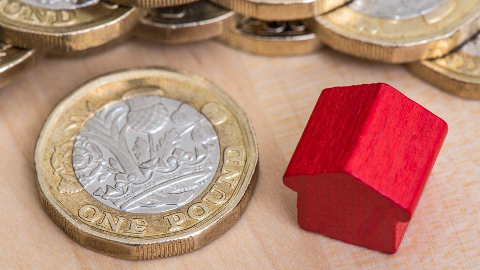 A red wooden model of a house next to a pile of UK pound coins