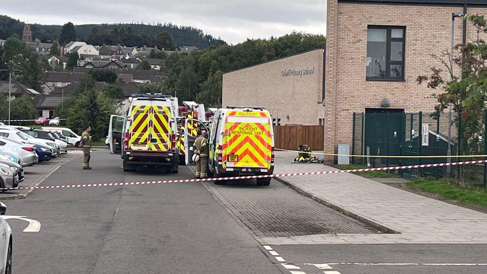 Emergency services vans in the school car park, with red and white tape sealing off the road. Fire crews are standing next to the vehicles.